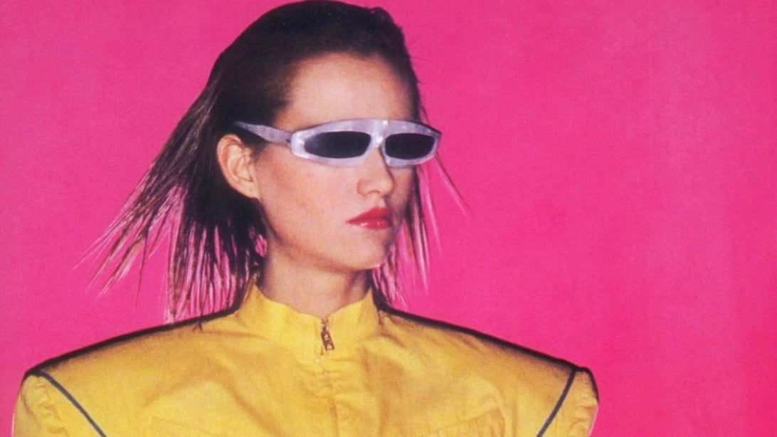 Person mit schmaler weißer Sonnenbrille und gelber Jacke vor pinkem Hintergrund